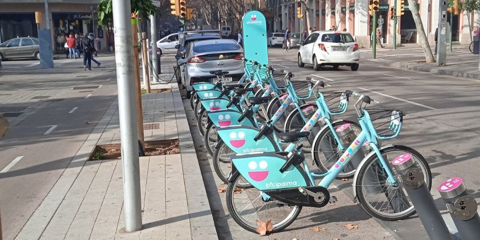 Foto d'una estació del sistema de bicicleta pública BiciPalma a la plaça Alexander Fleming amb carrer Balmes de Palma. A darrera, un cotxe elèctric endollat a un punt de càrrega. A l'esquerra, un carril bici sobre la voravia. Aquesta imatge serveix per a il·lustrar alternatives sostenibles en matèria de mobilitat i aparcament, com el bikeshare, el carsharing o cotxe compartit, una alternativa al rent a car o lloguer de cotxes convencional.